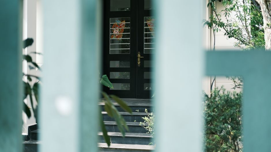 A rustic, rust-colored metal gate set within a lush, green garden hedge archway. The hedge is dense with vibrant, broad-leaved foliage, neatly trimmed to form a rounded top that frames the gate. Behin